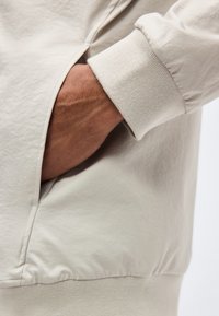 Χέρι που έχει εισαχθεί στην πλαϊνή τσέπη ενός ανοιχτό μπεζ μπουφάν με ριμπ μανσέτα και τελείωμα, δείχνοντας την υφή και τη εφαρμογή του ενδύματος.