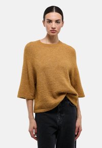 Femme aux cheveux foncés portant un pull en tricot à manches courtes couleur moutarde, rentrée dans un jean noir taille haute, debout devant un fond clair uni.