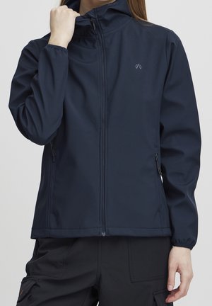 Persoon in een marineblauwe zip-up jas met een hoge kraag en zwarte cargobroek, die met één hand de kraag aanpast.