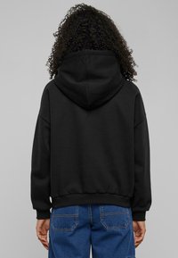 Svart hettegenser med ribbede ermer og hem. Stoffet ser mykt ut med en avslappet passform. Brukeren har på seg blå denimshorts.