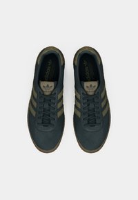 Σκούρα πράσινα sneakers από σουέτ με τρεις χακί ρίγες, καουτσούκ σόλα, στρογγυλή μύτη και σχέδιο με κορδόνια. Το λογότυπο είναι εμφανές στη γλώσσα και στη εσωτερική σόλα.