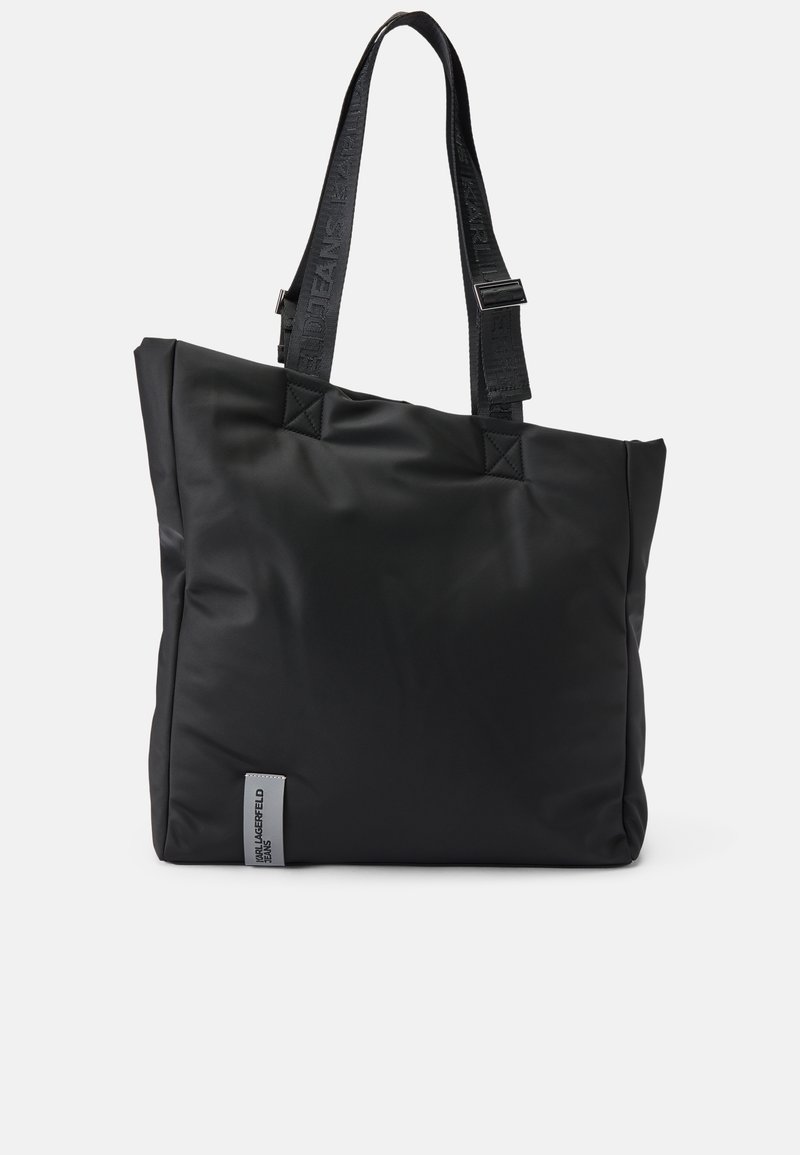 Saco tote de nylon preto com formato retangular, superfície texturizada e alças longas que apresentam o logotipo da marca. Inclui um pequeno detalhe em forma de etiqueta prateada.