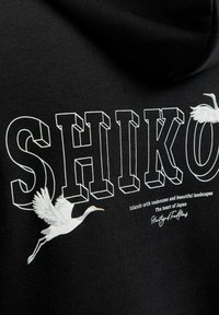 Czarny materiał z dużym, białym, trójwymiarowym napisem „SHIKO”, na którym znajdują się dwa lecące białe żurawie oraz mniejszy tekst dotyczący japońskich wysp i dziedzictwa.