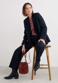 Marineblazer en -broek, kastanjebruin gestreept shirt, houdt een rode leren bucketbag vast; zwarte spitse laarzen; houten kruk.