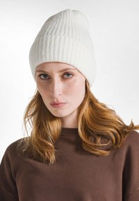 Valkoinen ribatut pipo, joka on valmistettu pehmeästä kankaasta, supistuvan huipun ja saumattoman muotoilun kanssa. Yhdistettynä ruskeaan sweatshirteeseen, se vaikuttaa mukavalta ja lämpimältä.