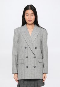 Jeune femme aux longs cheveux noirs portant un blazer gris surdimensionné à double boutonnage avec des rayures verticales et des boutons noirs sur une jupe plissée.