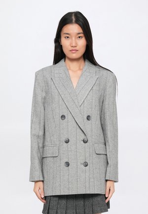 Noor naine, kellel on pikad tumedad juuksed, kannab suurust ületavat halli kahekordse nööbiga bleiserit, millel on vertikaalsed triibud ja mustad nööbid, ning pleegitatud seeliku alla.