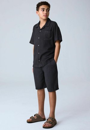 Teismeline poiss seisab, kandes musta lühikese varrukaga nööpidega särki, sobivaid lühikesi pükse ja pruune pandlaga sandaale, vaadates küljele käed taskutes.