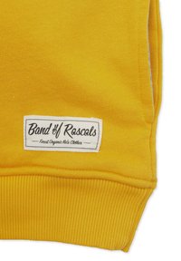 Keltaista ribbineulosta, jossa on valkoinen lähellä helmaa kiinni ommeltu etiketti, jossa lukee "Band of Rascals, Finest Organic Kids Clothes".