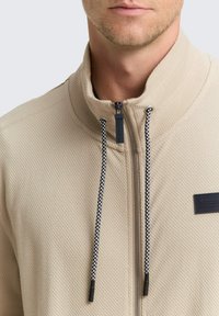 Giacca beige con zip, superficie testurizzata, dettagli con cordoncino nero e bianco, e un piccolo logo nero sul petto.