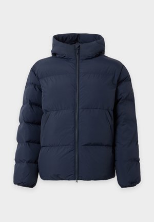 Un mannequin portant une doudoune bleu marine à capuche, un pantalon assorti et des baskets blanches se tient devant un fond de studio blanc uni.
