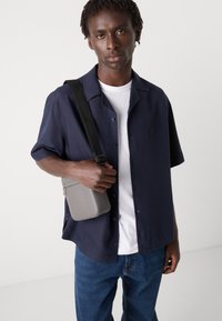 Sötétkék, rövid ujjú ing, amelyen egy zseb található, fehér póló fölé viselve, kék farmernadrággal párosítva, és egy szürke crossbody táska a kezében.