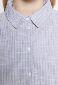 Camicia di lino azzurro chiaro con colletto, caratterizzata da un motivo a righe verticali testurizzate e bottoni bianchi lungo la parte frontale.