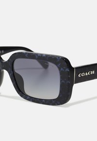 Juodos ir tamsiai mėlynos raštuotos saulės akinius su kvadratiniais rėmeliais, gradientiniais lęšiais ir metaliniu "COACH" logotipu ant šventyklų.