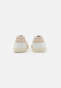Sneaker bianche in pelle con un tallone in suede beige. Presentano il logo "VEJA" in rilievo e una suola in gomma panna. Design pulito, accenti minimi.