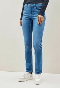 Light blue jeans made of denim with a slim fit, five-pocket design, and a button closure, paired with white sneakers on a neutral background.