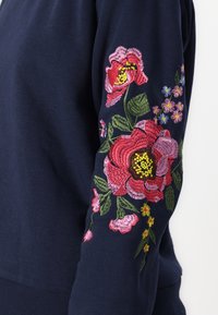 Manga de color azul marino con grandes flores rojas y rosas bordadas, hojas verdes y pequeños acentos florales amarillos y azules.