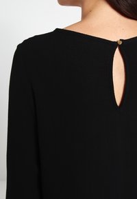 Robe noire à manches longues, avec une ouverture en forme de serrure au dos, agrémentée d'un petit bouton doré. Tissu texturé.