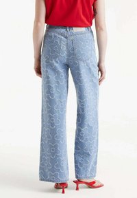 Jeans de mezclilla azul claro con pierna ancha, adornados con bordados florales blancos y bolsillos traseros. Combinados con una blusa roja y tacones rojos.
