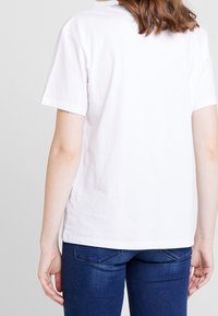 Camiseta blanca de algodón con mangas cortas, escote redondo y dobladillo recto. La parte trasera es lisa, sin patrones ni gráficos visibles.