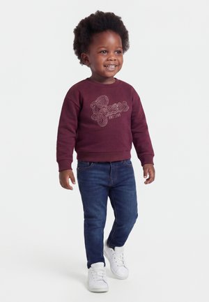 Tout-petit souriant marchant, portant un sweat-shirt bordeaux avec le texte "Guess", un jean bleu et des baskets blanches sur un fond blanc uni.
