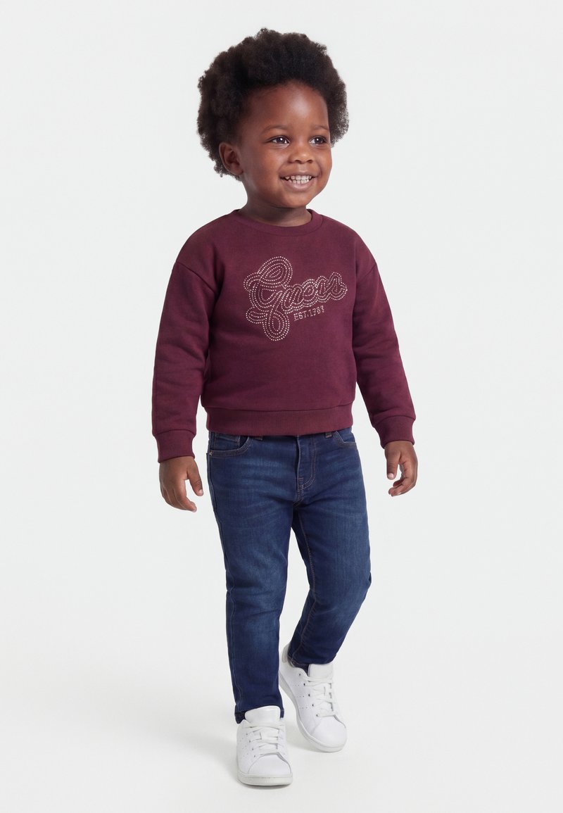 Tout-petit souriant marchant, portant un sweat-shirt bordeaux avec le texte "Guess", un jean bleu et des baskets blanches sur un fond blanc uni.
