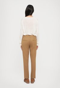 Femme aux longs cheveux noirs, portant un haut blanc à manches longues, un pantalon droit couleur camel et des chaussures compensées marron, se tenant de dos.