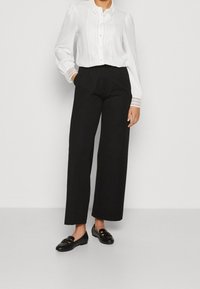 Chemisier blanc avec accents en dentelle et fermeture à boutons, associé à un pantalon noir ample et des mocassins noirs ornés d'une garniture décorative.