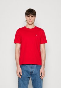 T-shirt en coton rouge avec un col rond et des manches courtes. Présente un petit logo sur la poitrine et un ourlet droit. Porté avec un jean en denim bleu.