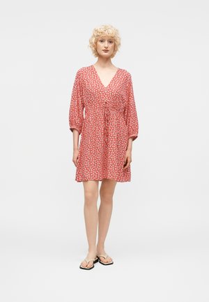 Γυναίκα με σγουρά ξανθά μαλλιά που φοράει κόκκινο φόρεμα με λουλούδια, V λαιμόκοψη και μανίκια τριών τετάρτων, και μπεζ σανδάλια, στέκεται μπροστά σε απλό φόντο.