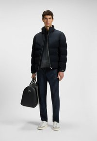 Donkerblauwe pufferjack met een hoge kraag en ritssluiting. Gepaard met gestreepte marineblauwe broek en een zwarte handtas. Witte sneakers met grijze accenten.