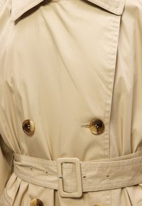 Trench-coat beige avec un large col, des boutons double boutonnage effet écaille et une ceinture assortie avec une boucle rectangulaire.