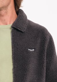 Chaqueta de forro polar gris con cuello y cremallera. Presenta el logo blanco "VOLCOM" en el lado izquierdo del pecho. Camiseta verde lisa debajo.