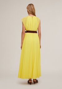 Abito maxi giallo plissettato con scollatura rotonda e maniche a cappuccio, cintrata in vita con una cintura marrone. Tessuto morbido e leggero.