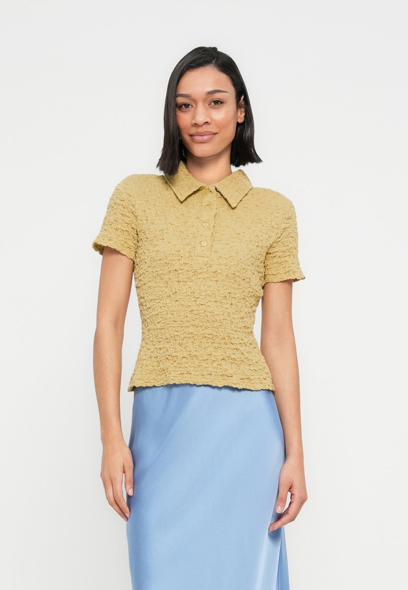 Γυναίκα με ίσια μαύρα μαλλιά, που φοράει μια κίτρινη πόλο μπλούζα με υφή και μια λεία ανοιχτό μπλε φούστα, στέκεται μπροστά σε λευκό φόντο.