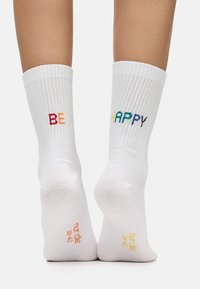 Valkoiset puuvillasukat, joissa on ribbireunukset, esittelevät punaisella värillä "BE" ja monivärisillä kirjaimilla "HAPPY". Pohjassa on teksti, joka sisältää tuotemerkinnän ja koon tiedot.