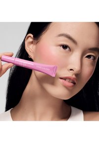 Różowa tubka produktu Estée Lauder trzymana blisko twarzy modelki, z gładką teksturą i wyraźnie widocznym logo. Modelka ma czystą cerę i czarne włosy.