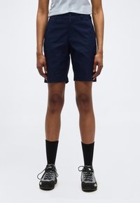 Άτομο που φοράει σορτς μήκους μέχρι το γόνατο σε ναυτικό μπλε, μαύρες κάλτσες μέχρι τη μέση της γάμπας και γκρι-μαύρα πεζοπορικά παπούτσια, στέκεται σε ένα απλό λευκό φόντο.