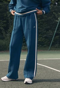 Türkisfarbene Jogginghosen mit weißen Seitenstreifen, die eine lockere Passform und weiche Textur bieten, kombiniert mit weißen Sportschuhen auf einem Sportplatz.