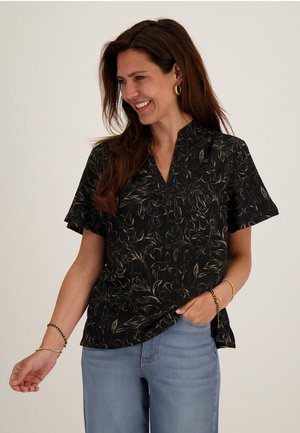 Vrouw glimlacht, draagt een zwarte korte mouwen blouse met bloemenprint en lichtblauwe spijkerbroek, met gouden creolen en armbanden, poseert tegen een effen achtergrond.