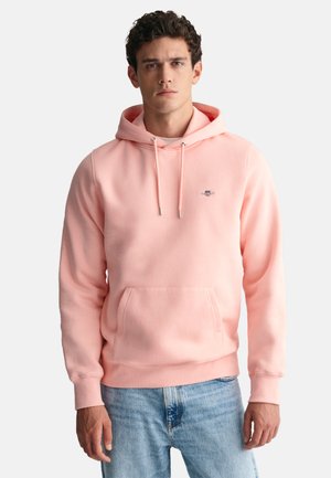 Giovane uomo che indossa una felpa con cappuccio rosa chiaro con tasca frontale e coulisse, abbinata a jeans azzurri.