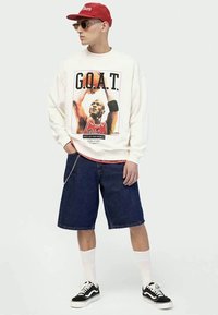 Uomo con cappello rosso, occhiali da sole, felpa bianca con stampa Michael Jordan G.O.A.T., pantaloni corti di denim, calze bianche e sneakers nere, in piedi con le mani in tasca.