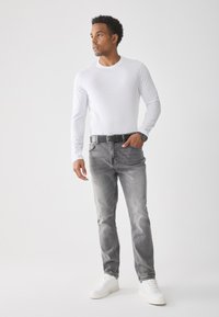 Weißes Langarmhemd, graue Jeans mit hellerem Waschung und Gürtel, weiße Sneakers, stehende Pose vor einem schlichten Hintergrund.