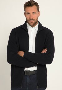 Marinblå ribbad cardigan med hög krage, stickad textur, dragkedja och förlängda muddar, bärs över en vit skjorta med krage.