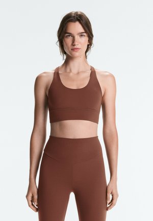 Femme debout face à l'avant, portant un soutien-gorge de sport marron et des leggings taille haute sur un fond clair uni.
