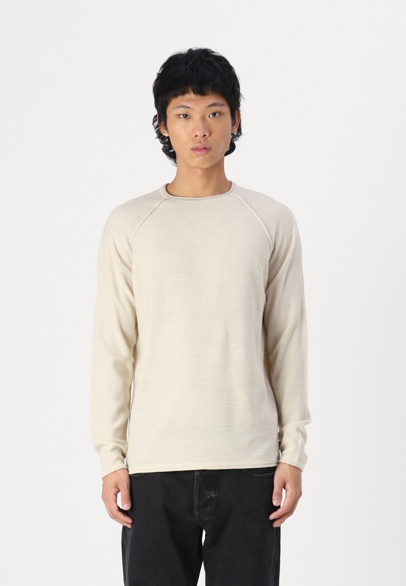 Chemise beige à manches longues avec un motif texturé, dotée d'un col rond et de manches raglan. Portée sur un pantalon noir, au design minimal.