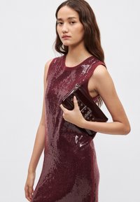 Vinovo červené šaty bez rukávov s flitrovou textúrou; modelka drží lesklé vinovo červené psaníčko s kovovými detailmi a zapínaním na zips.