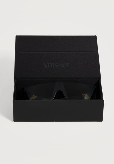 Lunettes de soleil noires Versace avec de larges verres foncés exposées dans un étui noir ouvert avec le logo Versace sur le couvercle.