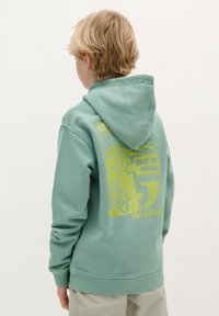Felpa verde menta con tasca a marsupio e grafiche gialle sulla schiena, tra cui un design di teschio e pezzo di puzzle. Materiale morbido, misto cotone.