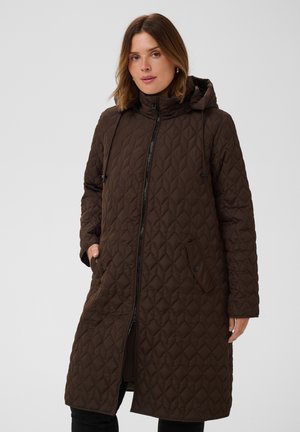 Femme portant un long manteau marron matelassé avec une capuche, debout avec une main dans la poche devant un fond clair uni.